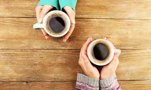 Two women sit across from each other at a wooden table, each holding a warm cup of coffee. The image evokes comfort, connection, and shared experience—reflecting the supportive nature of the group for mothers who have lost their mothers.